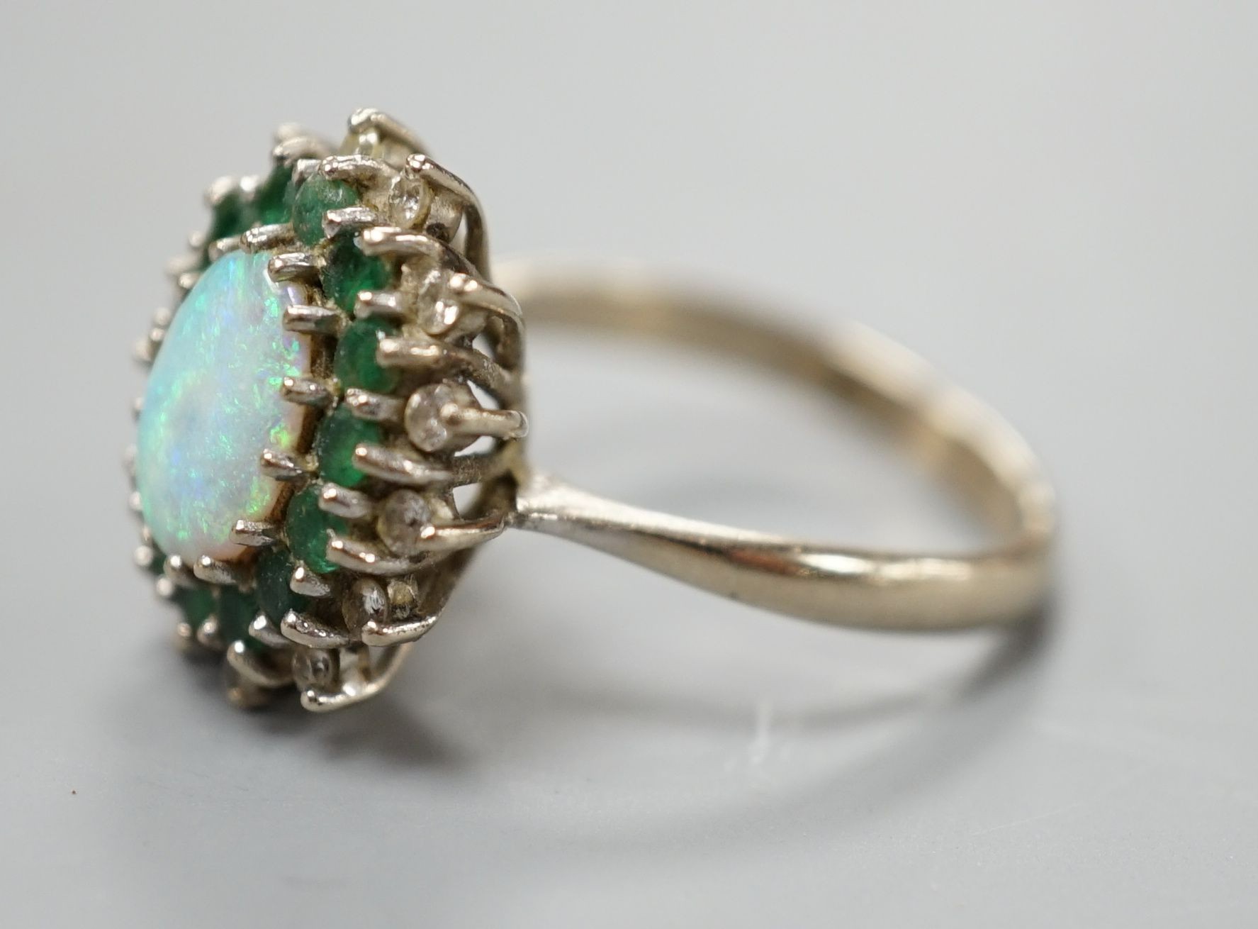 An 18ct white metal, white opal, emerald and diamond set oval cluster ring, size O, gross weight 6.2 grams.